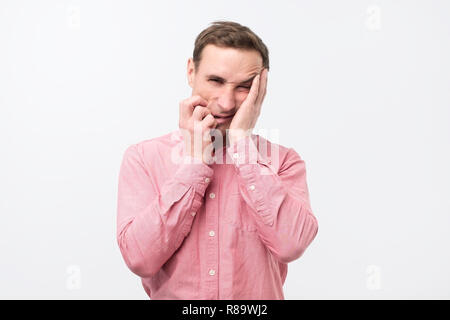 Gloomy man in pink shirt, holding hands on face, frowning and staring with helpless expression Stock Photo