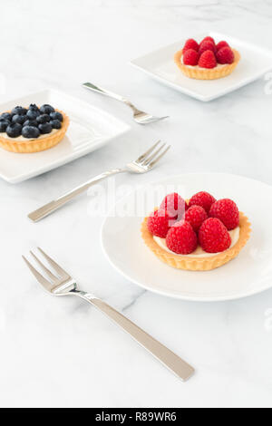 Raspberry and blueberry tartlets filled with vanilla cream on white porcelain plates and white marble. Stock Photo