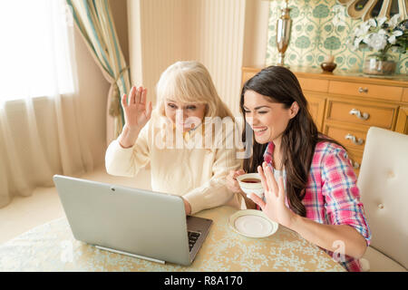 Two Lovely Women Wave To The Laptop Camera, Greeting A Friend. Smiling Daughter With A Cup Of Tea And Happy Senior Mother Wave To The Skype Caller. Stock Photo