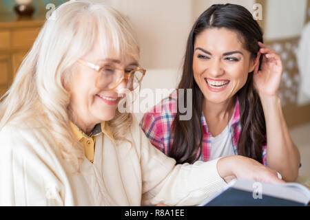 Two Wonderful Women Laugh Over Funny Story They Have Read From A Book. Daughter Looks With Love And Care On Her Senior Mother In Glasses Smiling. Stock Photo