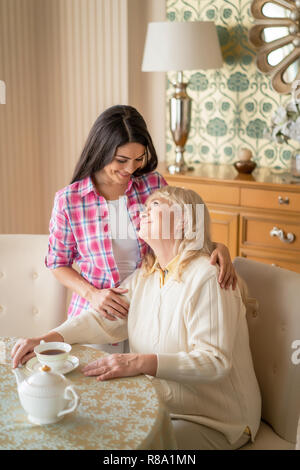 Elderly Woman Drinks Black Tea And Looks At Her Lovely Daughter Who Hugs Her Tightly Stock Photo