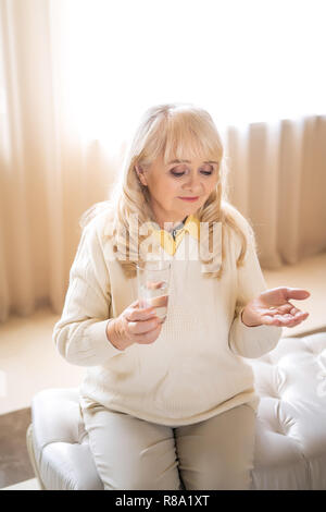 Tired Old Woman Looks Down On The Pills In One Hand While Holding A Glass Of Water In The Other Stock Photo