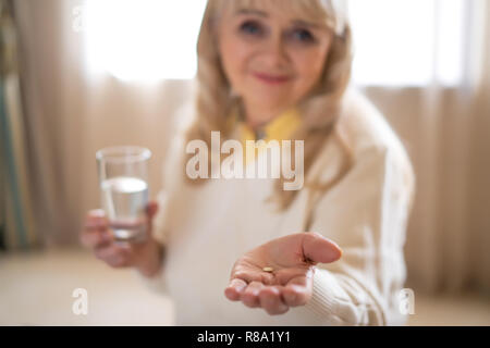 Blurred Smiling Senior Woman Shows A Pills In Her Palm While Holding A Glass Of Water. Close-up. Medication Stock Photo