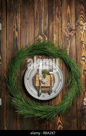 christmas table setting at brown wooden background with white plate, silver cutlery and paper envelope. xmas concept in pine circle. vertical top view Stock Photo