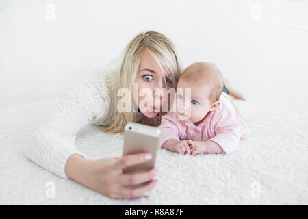 Funny baby girl make selfie on mobile phone and lying near her mother. Newborn looking at the camera and smiling. Stock Photo