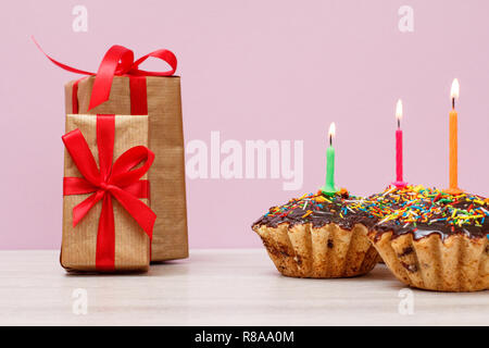 Gift boxes and three tasty birthday cupcakes with chocolate glaze and caramel, decorated with burning festive candles on lilac background. Happy birth Stock Photo