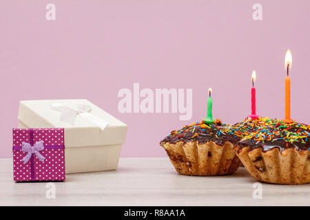 Gift boxes and three tasty birthday muffins with chocolate glaze and caramel, decorated with burning festive candles on lilac background. Happy birthd Stock Photo
