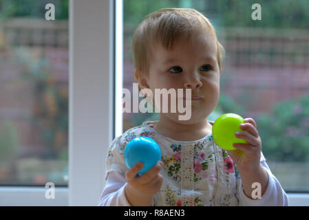 Few months old baby sitting and holding blue and green ball. Glass door and garden in the background. Stock Photo