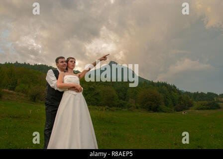 Freshly married couple embraced and bride dressed in white pointing her finger top right outside in front of the mountain in Slovakia Stock Photo