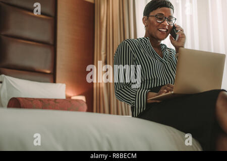Happy mature formal dressed woman talking on mobile phone while sitting in the hotel room using laptop. African businesswoman on tour working from hot Stock Photo