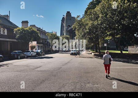 A women on her own walking along Lower Fort Street with the Sydney Harbour Bridge in the distance showing its size against buildings and cars Stock Photo