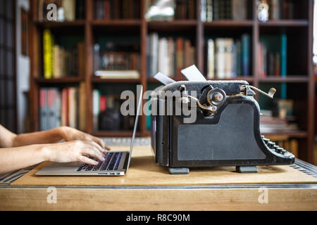 woman hands type on modern notebook. old vintage dust-covered typewriter with sheet of white paper near modern notebook on bookcase background. modern Stock Photo