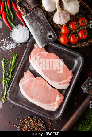 Raw pork loin chops in plastic tray with salt and pepper and vintage meat hatchets and hammer on rusty board.Red pepper, tomatoes and garlic. Stock Photo