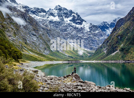 A couple of walkers looking out over Lake Marian in the Darran Mountains of Fjordland in South Island New Zealand Stock Photo