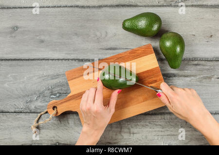 Tabletop view, woman hands holding chef knife, cutting avocado on chopping board, two whole avocados near on gray wood desk. Stock Photo
