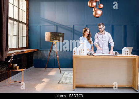 Young woman and man as an architects working with blueprints, laptop and house models in the office. Wide interior view Stock Photo