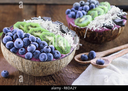 Açai smoothie bowl with fresh kiwi, frozen blueberries, organic coconut and dark chocolate pieces with wooden spoons served in coconut bowls over a ru Stock Photo