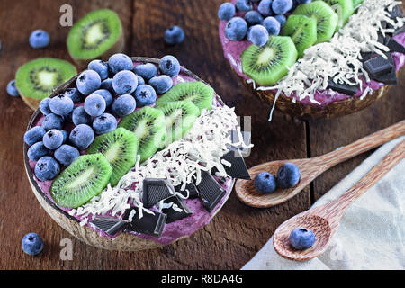 Açai smoothie bowl with fresh kiwi, frozen blueberries, organic coconut and dark chocolate pieces with wooden spoons served in coconut bowls over a ru Stock Photo