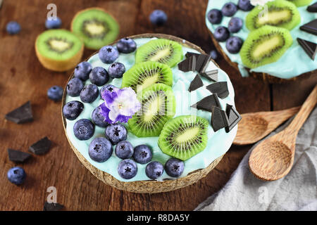 Blue spirulina and berry smoothie bowl, fresh blueberries, kiwi and chocolate pieces with wooden spoons served in coconut bowls over a rustic backgrou Stock Photo