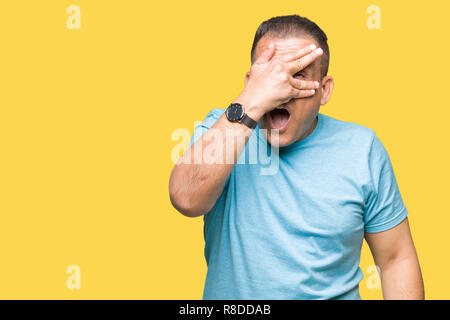 Middle age arab man wearing blue t-shirt over isolated background peeking in shock covering face and eyes with hand, looking through fingers with emba Stock Photo