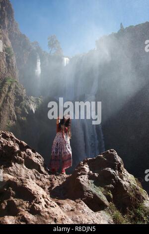 A young, hippie female tourist poses in front of a dramatic waterfall, making the peace sign and standing in a bright beam of light Stock Photo