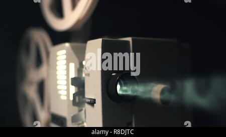 Front view of an old-fashioned antique Super 8mm film projector, projecting a beam of light in a dark room next to a stack of unraveled film reels Stock Photo