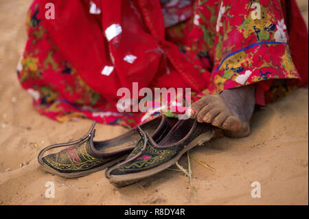 A gypsy Indian woman wearing a red sari is sitting barefoot in the sand after taking her shoes off, Thar Desert, Pushkar, Rajasthan, India. Stock Photo