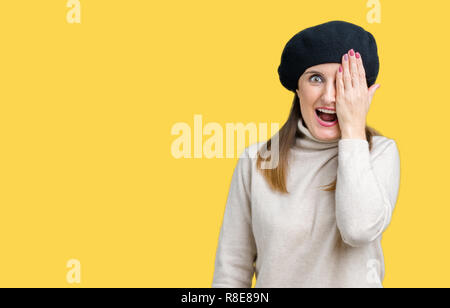 Middle age mature woman wearing winter sweater and beret over isolated background covering one eye with hand with confident smile on face and surprise Stock Photo