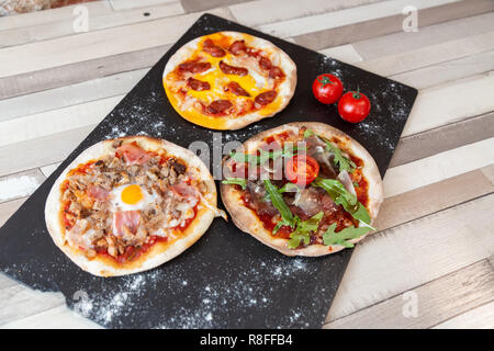 three different pizzas on a black board garnish with flour and two cherry tomato on a wooden white table Stock Photo