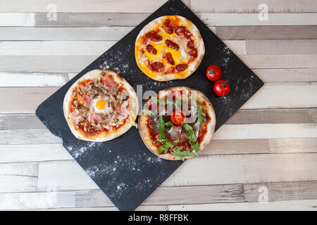 Aerial view of three different pizzas on a black board garnish with flour and two cherry tomato on a wooden white table Stock Photo