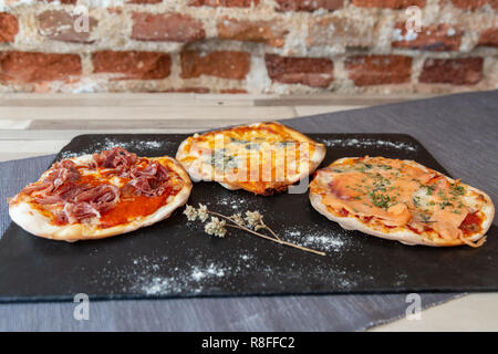 three different pizzas on a black board garnish with flour and dry flower on a wooden white table a gray tablecloth and a brick wall Stock Photo