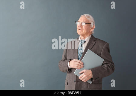 Senior man teacher wearing glasses studio standing isolated on gray holding book looking aside thoughtful Stock Photo