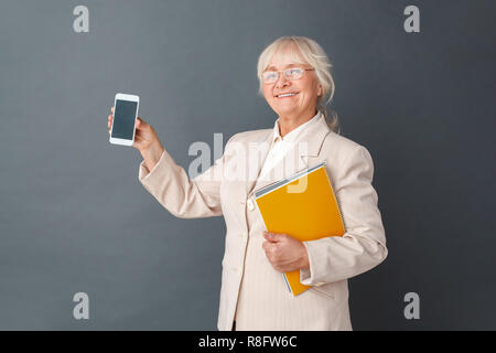 Senior woman in fromal suit and glasses studio standing isolated on gray holding journal howing smartphone screen smiling happy Stock Photo