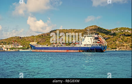 St. Barts - January 01, 2016: big cargo ship or barge blue color in French island bay of Saint Barth lemy with houses on green mountain, caribbean sea water and cloudy blue sky sunny summer vacation Stock Photo