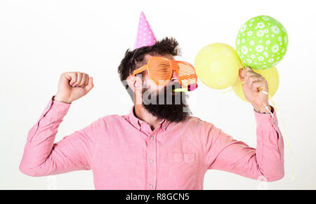 Celebration concept. Guy in party hat with air balloons celebrates. Hipster in giant sunglasses celebrating birthday. Man with beard and mustache on busy face blows into party horn, white background. Stock Photo