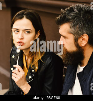 Woman and man with surprised faces have date at cafe. Patisserie and love concept. Couple in love takes a bite during break. Girl looks at bearded guy holding fork in mouth on brown terrace background Stock Photo