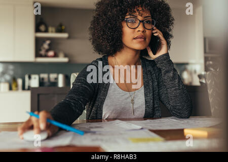 Woman with curly hair using desktop computer and talking on cell phone. Female architect working at her desk at home office. Stock Photo