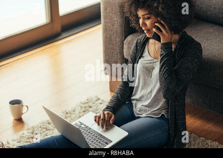 African woman sitting on floor using laptop and talking on cell phone. Female working using mobile phone and laptop at home. Stock Photo