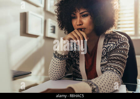 Thoughtful young woman looking away while drawing building blueprint sitting at table in home office. African female working on new floor plan at offi Stock Photo