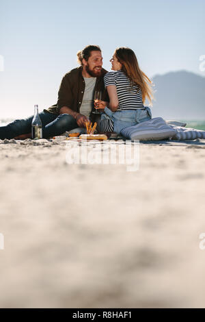 Beautiful couple enjoying in a good mood and picnic day on the beach. Low angle view of man and woman sitting by the beach with food and drinks. Stock Photo
