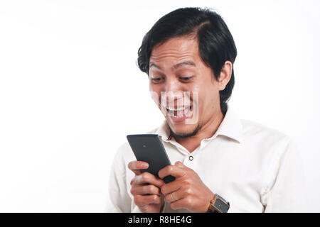 Photo image portrait of a funny young Asian man surprised and smiling while looking his smart phone. Holding phone with both hands while reading messa Stock Photo