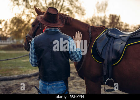 Brutal cowboy in jeans and leather jacket poses with horse on texas ranch, western. Vintage male person with animal, wild west Stock Photo