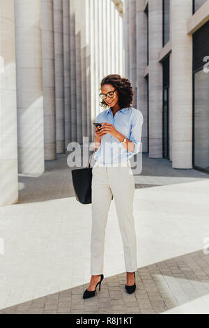 Businesswoman standing outside office building. Young professional using smartphone. Stock Photo