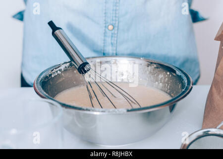 Close-up metal bowl with batter and whisk on white table, person wearing blue shirt standing on background. Stock Photo