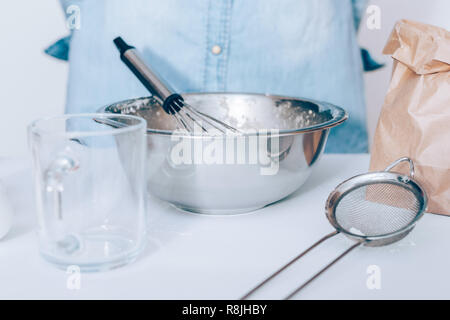 Close-up white kitchen table with utensils and metal bowl with batter and whisk, person wearing blue shirt standing on background. Stock Photo
