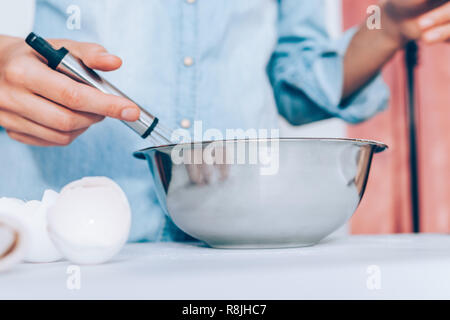 Candid image woman's hand whisking dough in metal bowl at white kitchen table, close-up. Stock Photo