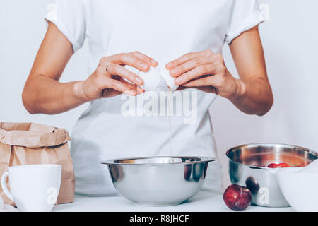 Female in white t-shirt breaking egg while cooking pie. Woman baker standing at table with metal bowl, fresh fruits, sack of flour, cup and preparing  Stock Photo