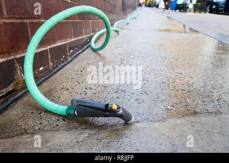 Low angel perspective of watering green hose and nozzle on a cement walkway used to clean pavement Urban city brick building Stock Photo