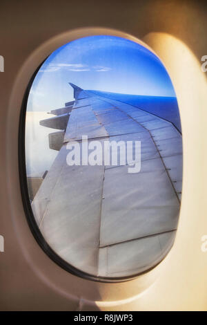 Oval of open viewing passenger window with surface of airplane wing and engines high in stratosphere during long distance flight in blue sky. Stock Photo