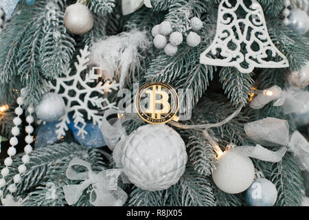 Gold coin of bitcoin lying on a white ball among the toys of the Christmas tree. Stock Photo
